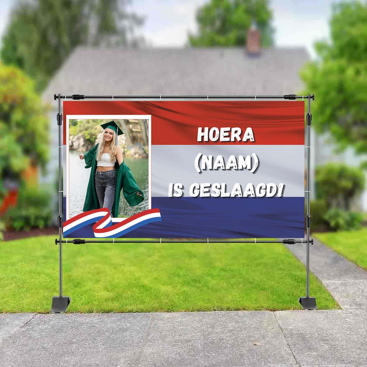 Spandoek met foto van geslaagde student in toga met diploma, Nederlandse vlag op de achtergrond en tekst 'Hoera (naam) is geslaagd!' in witte hoofdletters